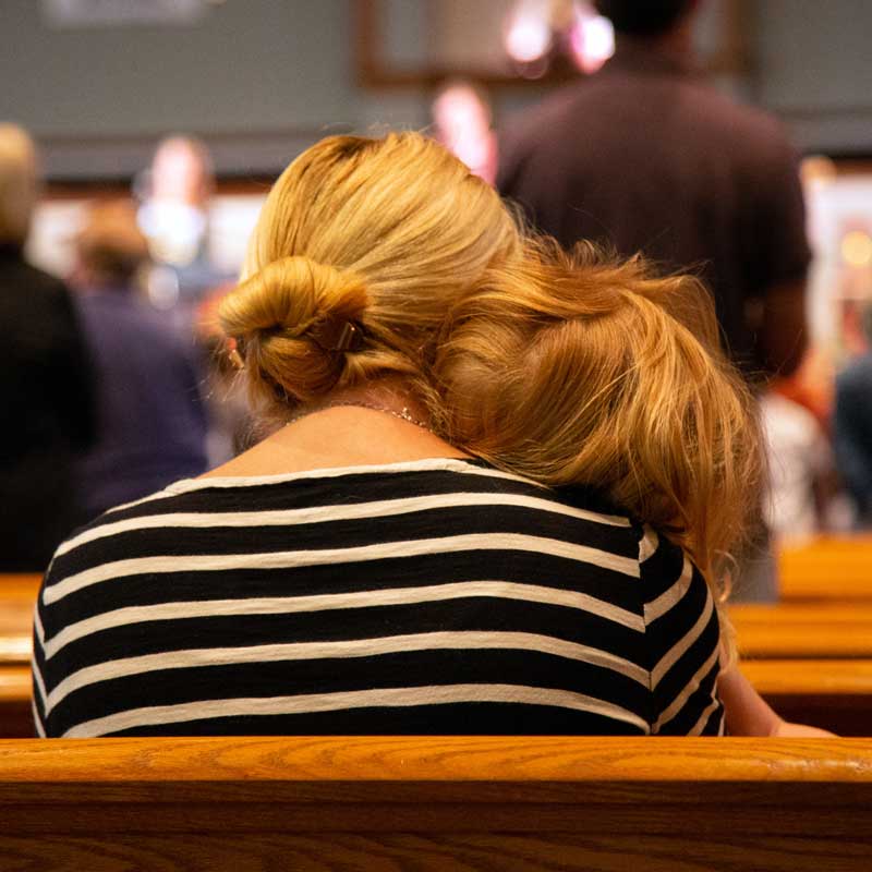 Taken from behind the pews, a mother is shown holding her young daughter on her shoulder while the worship team leads on stage.