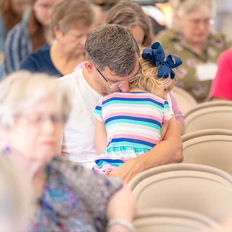 Taken from behind the pews, a mother is shown holding her young daughter on her shoulder while the worship team leads on stage.